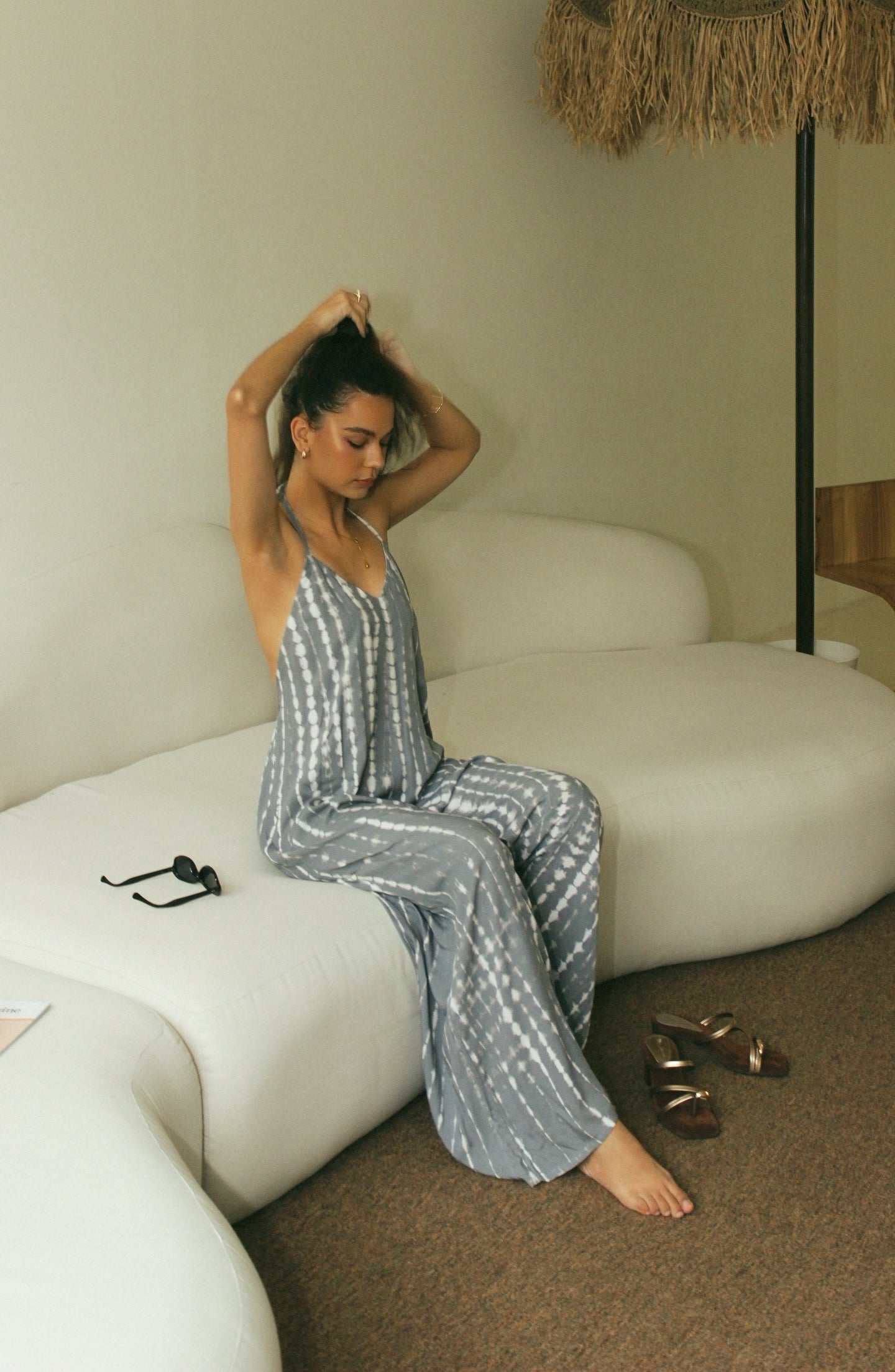 Woman in a bali tie-dye grey maxi jumpsuit sitting on a white couch in a cozy room.
